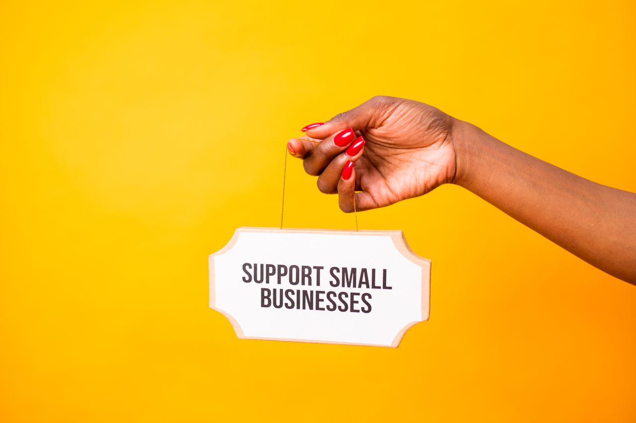 The Art of Drawing Readers In: Your attractive post title goes here Hand holding a 'Support Small Businesses' sign against a vibrant yellow background, promoting local commerce.